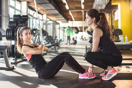 Téléchargez les photos : Jeune femme avec l'aide de son amie qui fait des redressements assis dans la salle de gym - en image libre de droit