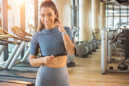 Photo for Young sporty woman putting earphone in ear to listen to the music while working out in gym. Attractive smiling girl. - Royalty Free Image