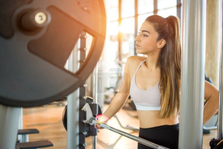 Photo for Young woman in sportswear leaning on barbell at gym. Attractive fitness girl in sportswear leaning on barbell in gym. - Royalty Free Image