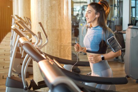 Beautiful young sporty girl wearing sportswear and ponytail and running on treadmill at gym. Fitness woman listening to music while running on treadmill.
