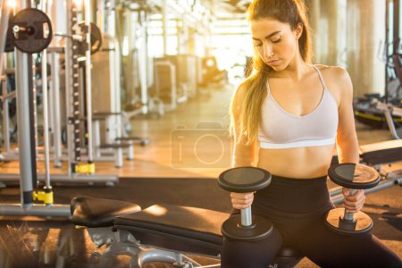 Photo for Young attractive athletic woman in sportswear sitting on a bench and holding dumbbells at gym - Royalty Free Image