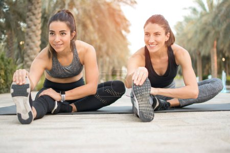 Dos mujeres de fitness sonrientes estirándose juntas en una colchoneta de ejercicio en el parque. Ajuste amigos niñas tocando los dedos de los pies durante el entrenamiento de fitness al aire libre