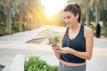 Photo for Young fitness woman with earphone using smartphone in the park - Royalty Free Image