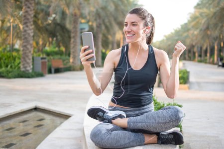 Téléchargez les photos : Femme de fitness excitée portant des écouteurs en utilisant le téléphone tout en étant assis dans le parc après avoir couru - en image libre de droit