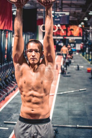 Téléchargez les photos : Fitness beau jeune homme torse nu faisant des exercices de trempage en utilisant des anneaux dans la salle de gym - en image libre de droit