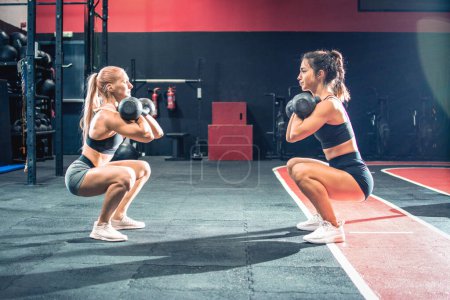 Téléchargez les photos : Deux jolies filles en position accroupie debout face à face soulevant une cloche de bouilloire pendant le cours d'exercice dans le gymnase - en image libre de droit