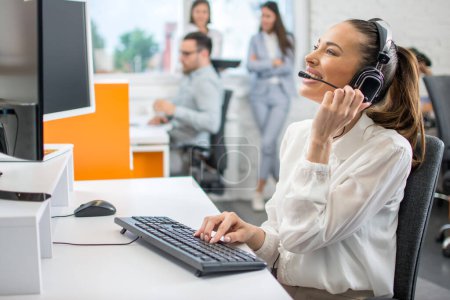 Téléchargez les photos : Sympathique opérateur de centre d'appels dame dans casque sans fil parler avec le client au bureau - en image libre de droit