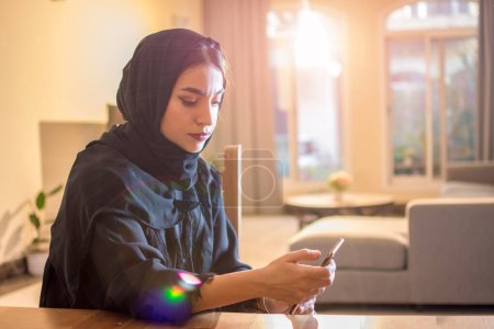Téléchargez les photos : Belle femme musulmane portant hijab en utilisant un téléphone intelligent tout en étant assis à la maison - en image libre de droit