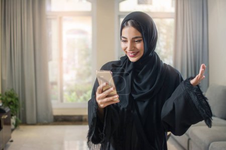 Téléchargez les photos : Portrait de jeune femme musulmane joyeuse en abaya en utilisant un smartphone tout en se tenant debout dans le salon - en image libre de droit