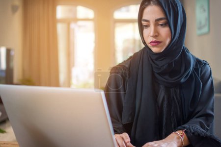 Téléchargez les photos : Jeune femme musulmane portant abaya en utilisant un ordinateur portable à la maison - en image libre de droit