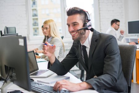 Téléchargez les photos : Homme d'affaires souriant avec casque de travail sur ordinateur de bureau dans le centre de support client - en image libre de droit