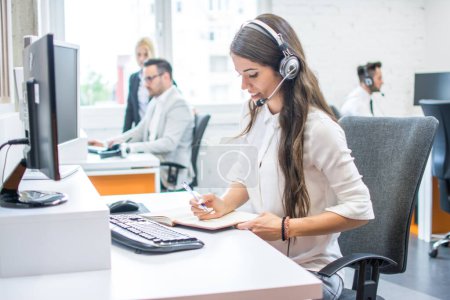 Photo for Side view of beautiful young lady working in office. Customer support worker taking notes while talking with client via headset at office desk. - Royalty Free Image