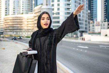 Foto de Hermosa mujer árabe en Abaya con bolsas de compras, teléfono y billetera enganchando un taxi en la calle - Imagen libre de derechos