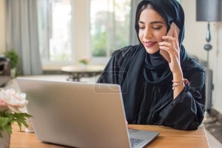 Téléchargez les photos : Belle jeune femme arabe du Moyen-Orient parlant sur un téléphone mobile et utilisant un ordinateur portable à la maison - en image libre de droit