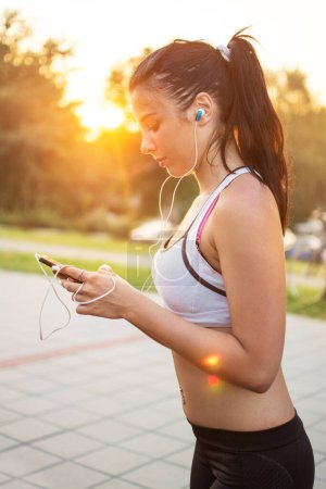 Fitness woman taking a workout rest for texting on her smartphone. She is listening to music outdoors