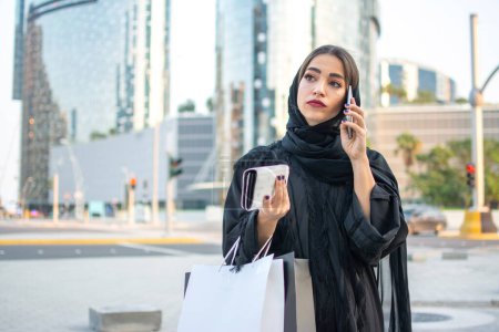 Foto de Mujer árabe en abaya tradicional llevar sosteniendo bolsas de compras y billetera en las manos y hablando por teléfono móvil mientras camina por la calle - Imagen libre de derechos