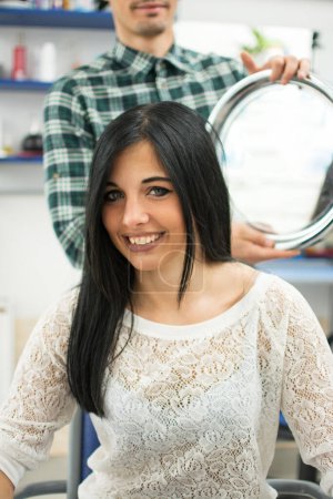 Portrait of smiling female customer. Young woman is looking her brand new hairdo while the hairdresser holding mirror