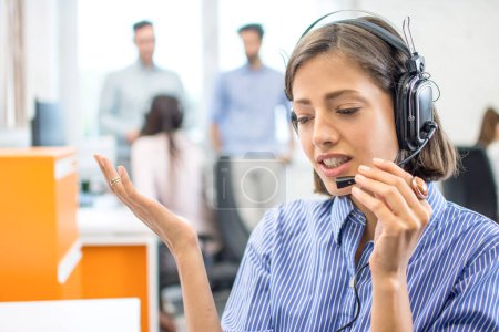 Foto de Retrato de cerca de una hermosa trabajadora del centro de llamadas usando auriculares manos libres y hablando con un cliente en la oficina - Imagen libre de derechos