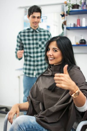 Téléchargez les photos : Jeune femme montrant pouce dans le salon de coiffure. Le coiffeur se tient derrière elle. - en image libre de droit