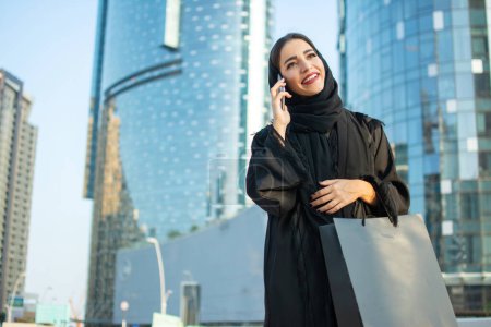 Foto de Mujer árabe feliz con bolsa de compras hablando en el teléfono móvil en la calle - Imagen libre de derechos