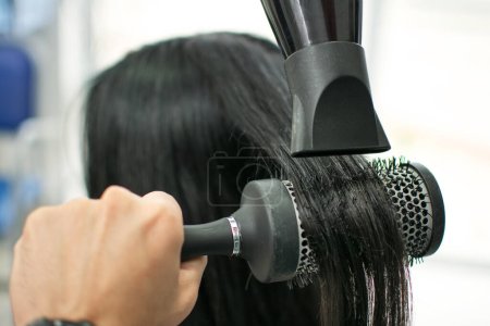 Foto de Primer plano de peluqueros manos secado pelo largo negro con secador de pelo y cepillo redondo - Imagen libre de derechos