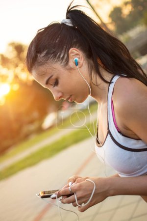 Téléchargez les photos : Jeune femme sportive utilisant un téléphone intelligent et écoutant de la musique en plein air au coucher du soleil - en image libre de droit