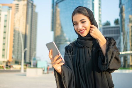 Téléchargez les photos : Belle jeune femme arabe souriante en vêtements traditionnels abaya à l'aide d'un smartphone dans la rue - en image libre de droit