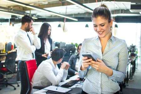 Téléchargez les photos : Femme d'affaires joyeuse utilisant smartphone au bureau tandis que ses collègues ayant réunion d'affaires en arrière-plan - en image libre de droit
