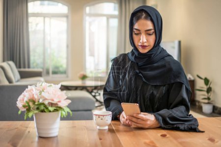 Photo for Beautiful muslim woman wearing abaya using smart phone while sitting at her luxury apartment - Royalty Free Image