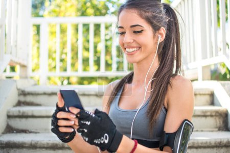Photo for Smiling sporty girl using phone during exercise break outdoors - Royalty Free Image
