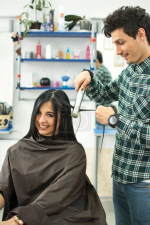 Foto de Joven estilista masculino usando una plancha de pelo en una mujer en el salón - Imagen libre de derechos