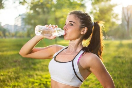 Foto de Joven mujer sonriente bebiendo agua de la botella después de ejercicio deportivo al aire libre al atardecer - Imagen libre de derechos