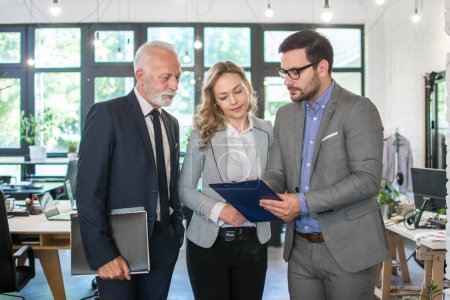 Photo for Business people working together in office. Serious senior businessman reading document with his two younger colleagues at team meeting - Royalty Free Image