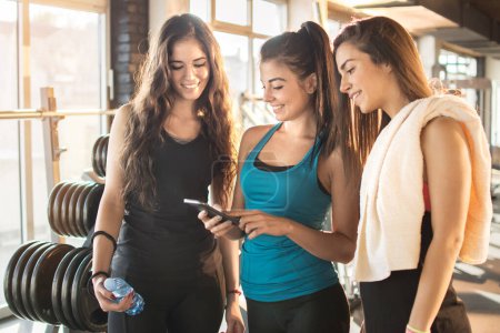Photo for Group of sporty female friends using app on mobile phone, looking at screen of smart phone, texting, watching photos while resting after practice, on break between exercises in gym. - Royalty Free Image