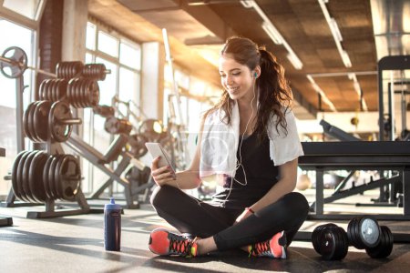 Foto de Joven mujer deportiva escuchando música en el teléfono inteligente en el gimnasio. Romper después del entrenamiento duro - Imagen libre de derechos
