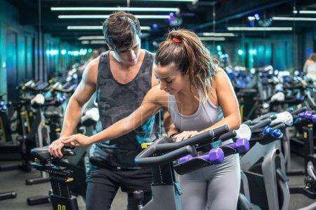 Photo for Young attractive woman with handsome trainer setting up exercise bike before cycling workout in gym - Royalty Free Image