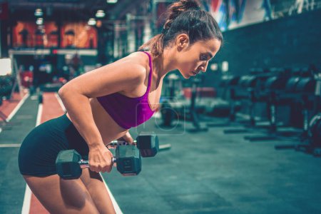 Photo for Young sportswoman in deadlift position exercising with dumbbells in the gym - Royalty Free Image