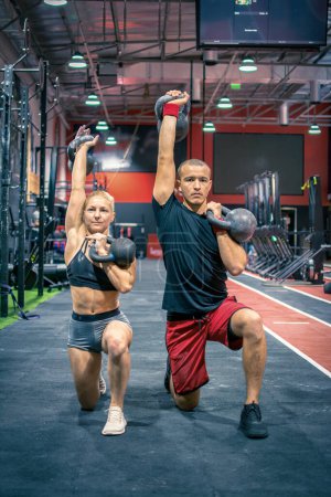 Téléchargez les photos : Vue horizontale du couple sportif faisant de l'exercice ensemble en position de fentes cloche de bouilloire de levage avec un bras levé et un près de la poitrine à la salle de gym - en image libre de droit