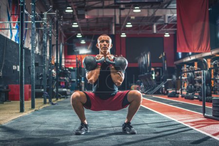 Téléchargez les photos : S'adapter homme en vêtements de sport faire des squats avec des poids lors d'un renforcement de l'entraînement à la salle de gym - en image libre de droit