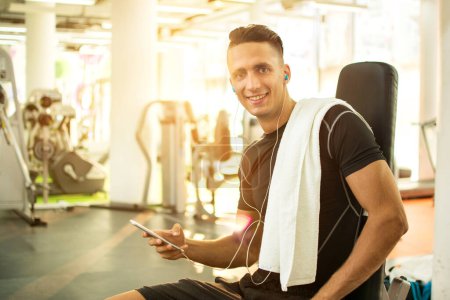 Téléchargez les photos : Beau jeune homme avec écouteurs utilisant un smartphone dans une salle de gym - en image libre de droit