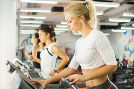 Photo for Young female athlete exercising on treadmill in a gym. She is setting up difficulty level on gym machine. - Royalty Free Image