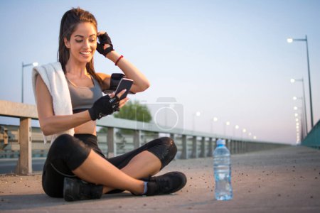 Téléchargez les photos : Jeune femme sportive utilisant le téléphone tout en s'asseyant sur le trottoir et se reposant après avoir couru sur le pont - en image libre de droit