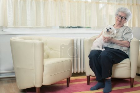 Foto de Senior mujer sosteniendo perro en su regazo y relajarse en la sala de estar. - Imagen libre de derechos