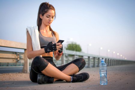 Téléchargez les photos : Jeune femme sportive assise sur le trottoir et utilisant le téléphone tout en faisant une pause après avoir couru à l'extérieur - en image libre de droit
