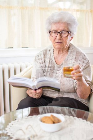 Seniorin liest Buch und trinkt Tee oder Apfelsaft.