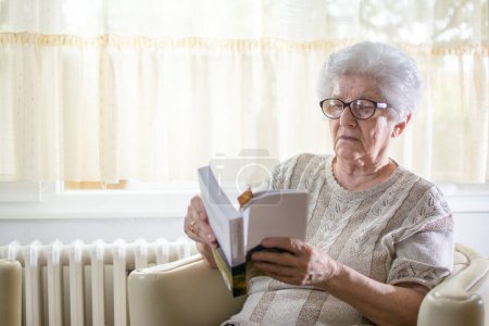 Foto de Mujer mayor leyendo libro mientras se relaja en el sillón en casa
. - Imagen libre de derechos