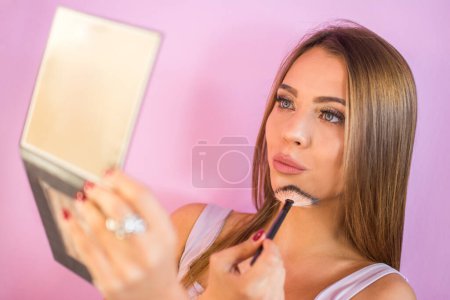 Photo for Young teenage girl applying make up on her face using a brush while holding a mirror. - Royalty Free Image