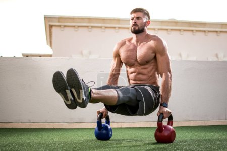 Photo for Muscular shirtless man workout with kettlebells in L Sit position outdoors. - Royalty Free Image
