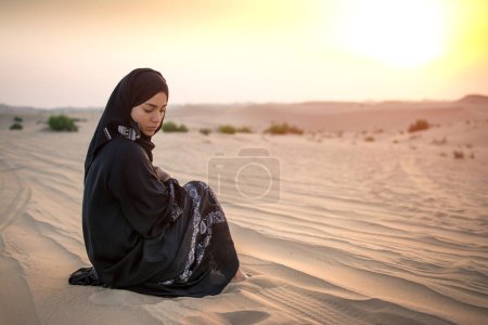 Foto de Mujer joven con ropa negra tradicional sentada en la arena contra el atardecer sobre el desierto. - Imagen libre de derechos