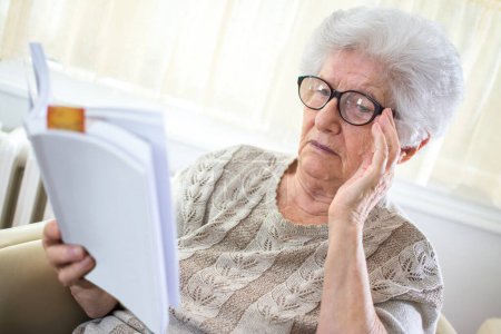 Foto de Mujer de pelo gris senior con anteojos leyendo libro en casa. - Imagen libre de derechos